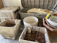 Photo of multiple large gardening pots including square and rectangular shapes, filled with dried plant materials, positioned beside a large gray plastic storage bin with lid containing folded tarp and twine.
