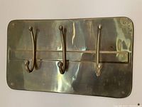 Front view of brass wall mounted coat hooks rack showing three double hooks affixed to a brass rectangular plate with rounded corners.