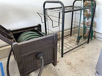 Black metal garden tool rack standing next to black plastic hose storage container with reel and green metal trellis pieces.