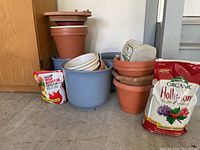 Photo showing a variety of medium outdoor gardening pots in terracotta and blue, with plastic plant trays, and two bags of organic gardening fertilizer.