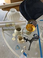 Photo of assorted household items including three rolls of wallpaper, decorative pitchers, vases, two blue seat cushions, some kitchen knobs and small candles placed on a glass table.