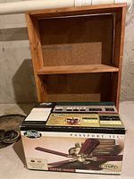 Boxed Hunter Passport III 52 inch ceiling fan with antique brass finish and wooden blades, new in box, placed in front of a small wooden 2-shelf bookcase.