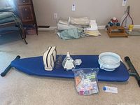 Blue ironing board flat on carpeted floor with accessories arranged on it: travel bag, timer, glass cup, stopper bowl, plastic bag with items, and iron cord.