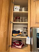 Open kitchen cabinet with multiple shelves showing the items: potholders on lower shelf, pitchers, measuring cups, sifter, apple peeler box, and grinder.
