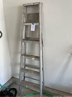Full view of 5-step aluminum folding ladder leaning against a wall, showing its overall size and condition with visible paint splatters and signs of use.
