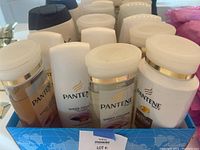 Image of multiple bottles of Pantene Pro-V hair care products with white and gold design, including shampoo and conditioner bottles.