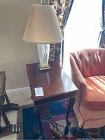 Wood side table with glass base lamp with pleated white lampshade beside an upholstered chair.