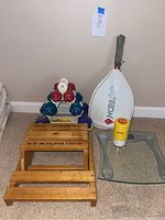 Photo showing all items in the lot: three sets of dumbbells stacked on a rack, white Everlast racquet with cover, yellow retro scale, wooden step stool, and glass-top side table.