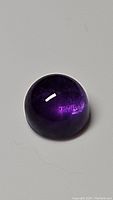 Close-up photo of deep purple round cabochon amethyst gemstone on a plain background, showing smooth polished surface and rich color.