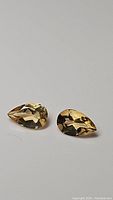 Close-up photo of a pair of pear-shaped golden citrine gemstones laying flat on a light surface, showing faceted cuts and clarity.