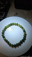 Top-down view of a green jade beaded stretchy bracelet arranged in a circle on a white circular surface with a dark background.