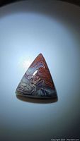 Photo showing a triangular fancy polished cabochon gemstone with red, white, gray and black layered lace patterning of natural crazy lace agate.