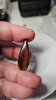 Close-up front view of the natural red tiger's eye pear cabochon held between fingers, showing wood-like chatoyant pattern and glossy surface.