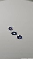 Three oval cut purple-blue natural tanzanite gemstones arranged in a row on a gray background.