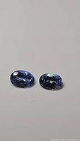 Photo showing two oval-cut tanzanite gemstones with purple-blue hue, placed side by side on white surface.