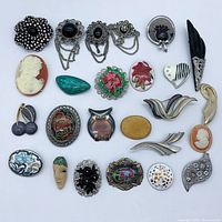 Photo shows full layout of 22 assorted brooches including flower shapes, cameos, enamel floral designs, heart and owl motifs, and decorative chain brooches in silver, gold and black tones.