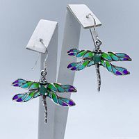 Pair of dragonfly dangle earrings shown hanging on white earring display stands, front view showing colorful enamel wings with green emerald center stones and marcasite accents on silver metal.