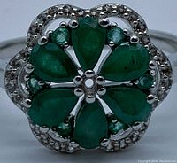 Close-up front view of ring showing six pear-shaped emeralds forming a flower surrounded by a halo of small round topaz stones in 14K white gold plated 925 sterling silver.