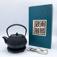 Full view of the mini iron cast tea kettle with original green box and papers beside it on a white background.