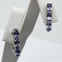 Pair of 3-stone oval tanzanite earrings on white earring display, front angle showing color and setting.