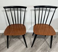 Two Mid-Century Modern Danish spindle back chairs with teak seats and black legs, positioned side by side on a wooden floor against a white wall.