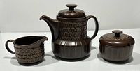Photo showing full set of dark brown Wedgwood Pennine teapot, creamer, and covered sugar bowl with textured geometric patterns on a white background.