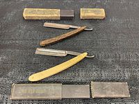 Two straight razors lying on a surface with one in its original box labeled 'THE MAB RAZOR REG.' and the other with a wood-grain handle placed nearby alongside two small rectangular containers.