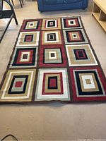 Top down photo of a rectangular area rug laid on the floor showing geometric multi-colored squares pattern in red, beige, brown, black, cream and gold tones in a 3x5 grid.