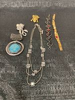 Photo showing the six pieces of costume jewelry laid flat on black textured fabric, including necklaces, a cat-shaped pin, a pearl bow pin, and two distinct bracelets.