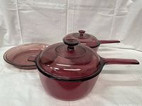Three-piece cranberry-colored glass cookware set including two lidded saucepans and one round baking dish on white fabric background.