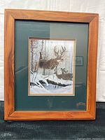 Framed artwork showing a buck deer leaping in a snow-covered forest with two other deer in the background, encased in a wooden frame with green matte.