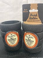 Front view of two tire-shaped drink holders with tread pattern and 'No. 1 Grandpa' label, stacked in black fabric background.