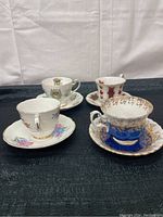 Four fine china teacups and saucers arranged on a dark surface, showcasing diverse floral and decorative patterns including blue and gold, floral house design, and red floral ribbons.