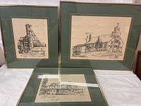 Three framed ink drawings displayed together on white fabric showing the industrial and architectural scenes.