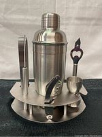 Front view of entire bar caddy set showing cocktail shaker, ice tongs, opener, stopper, jigger and caddy stand.