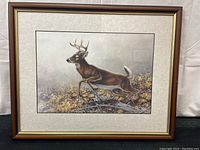 Full view of the framed Michael Dumas print showing the deer in jumping motion and the surrounding natural environment with leaves.