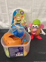 Mr. Potato Head figure standing with green hat, pink ears, white hands, red shoes, and various facial features. Plastic container next to figure containing extra parts and accessories used for the toy.