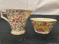 Pair of ceramic creamer and bowl featuring black floral outlines, colorful cross-stitch style flower decorations, and gold trim on a cream background.
