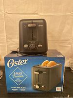Front view of black Oster 2-slice toaster placed on top of its original blue box.
