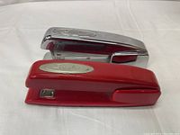 Photo showing one silver chrome and one red Swingline stapler on a white surface from a side angle.