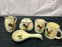 Photo showing four ceramic kitchenware items (pitcher, lidded sugar bowl, spoon rest, and mug) all cream-colored with blue trim and rooster illustrations.