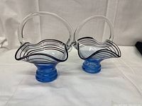 Front view of both art glass baskets showing blue base, brown stripes on clear wavy rim, and clear handles