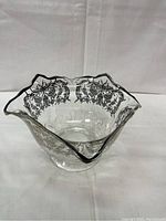 Side angle showing ruffled square shape, clear glass, and silver embossed floral designs on the rim and edges.