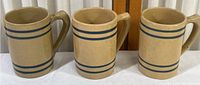 Three beige stoneware mugs with blue stripes, side by side, showing fronts and handles.