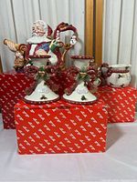 Front view of the Fitz and Floyd Christmas collection including Santa teapot, candlesticks, sugar dish on red patterned boxes.