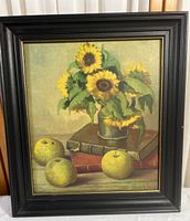 Framed still life print showing sunflowers in a metal container placed on books with three apples in the foreground in a black frame.