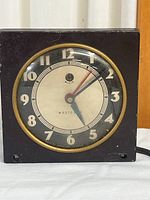 Square vintage Westclox travel clock with black casing and gold ring around face, showing time and condition.