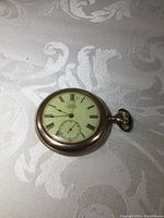 Angle view of the full pocket watch showing side and front with winding crown and bow, placed on a patterned white cloth.