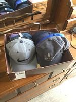 Photo showing 10 used ball caps in a small box on wooden dresser. Caps include Nike and other brands. Minor wear visible.