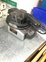 Photo of the Black & Decker belt sander showing the full unit with power cord wrapped around the handle, placed on a metal work surface.